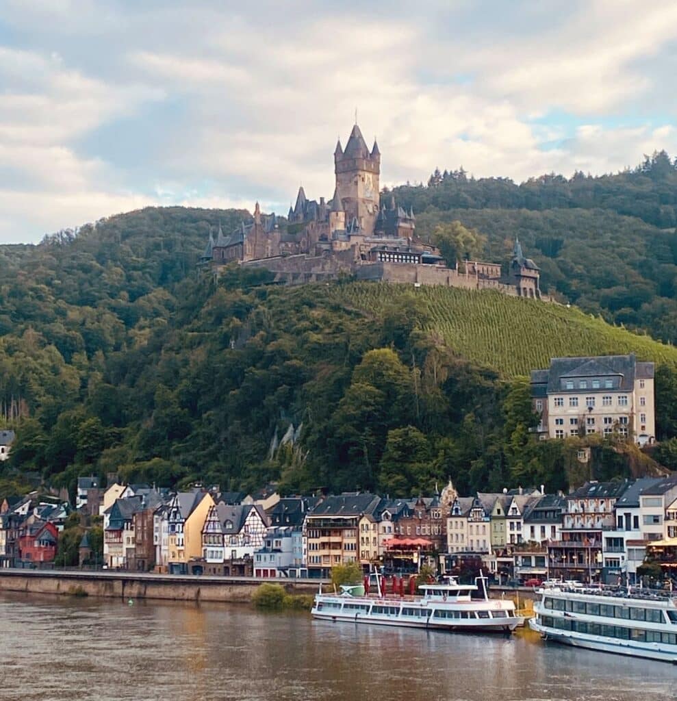 houses and boats on the river with a castle on the hill in the background